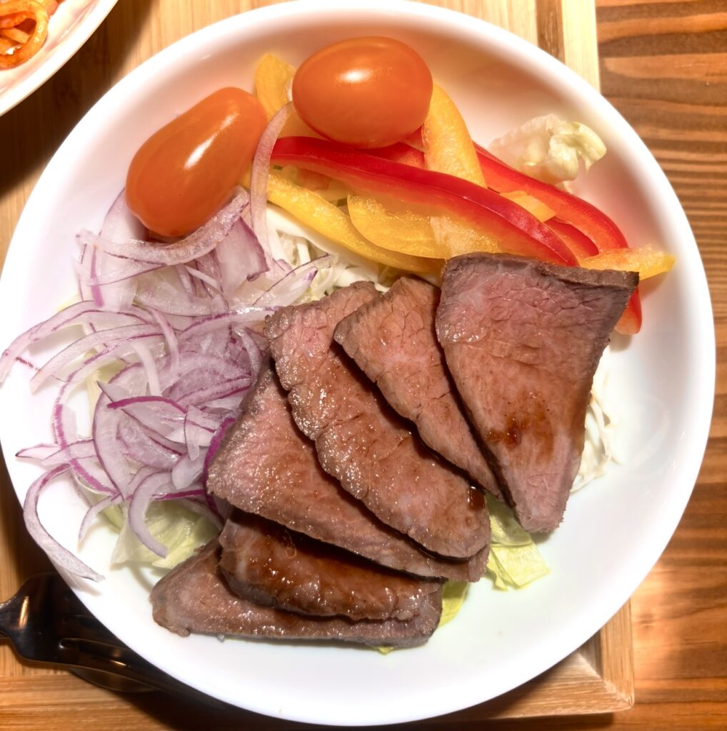 白い器に盛られたローストビーフサラダ。薄切りのローストビーフが重ねられ、その周りに紫玉ねぎスライス、赤と黄色のパプリカ、ミニトマト、レタスが彩りよく添えられている。色鮮やかで食欲をそそる一皿。