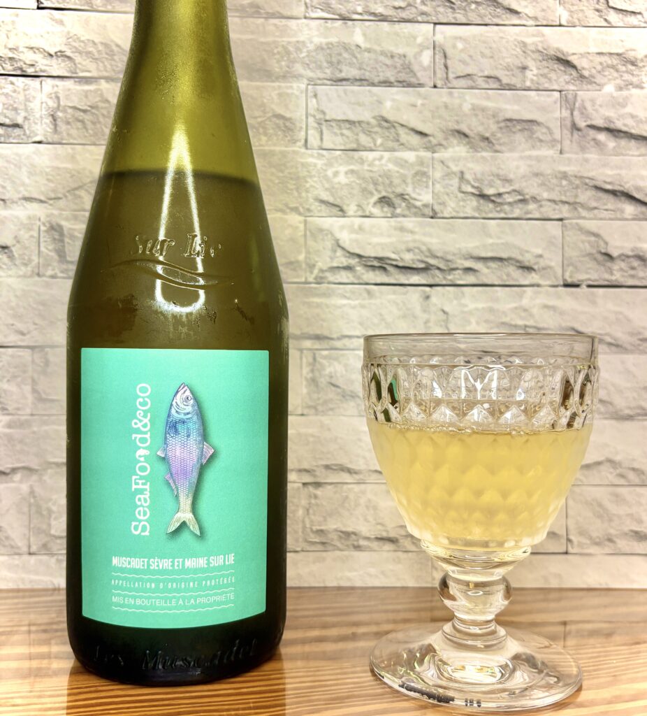 魚のイラストが描かれた緑色ラベルのワインボトルと、淡い黄色の飲み物が入ったカットグラスが木のテーブルに並んでいる」