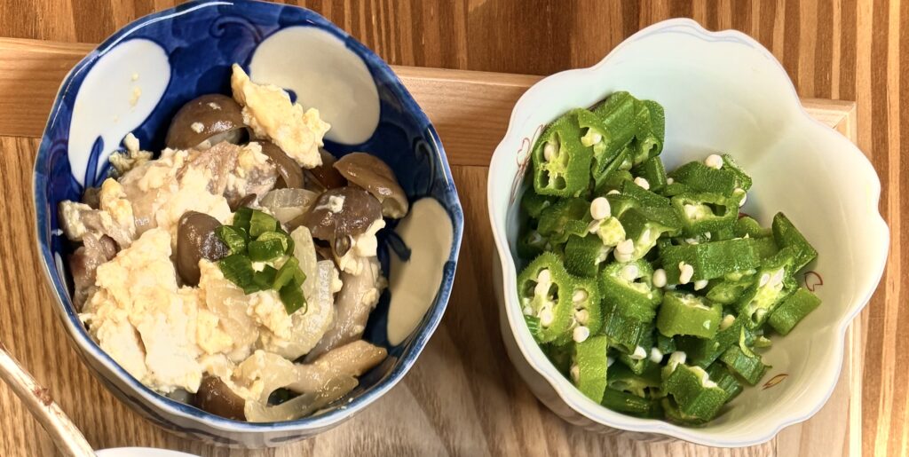 青い柄の小鉢には、しめじやしいたけなどのきのこと豆腐を炒め合わせたチャンプルーが盛られている。淡い黄色の卵と豆腐の柔らかな色合いに、茶色のきのこがアクセント。隣の白い小鉢には、鮮やかな緑色のオクラが小口切りで盛られ、粘りが光る副菜として並んでいる。彩りと栄養のバランスが取れた家庭の副菜セット
