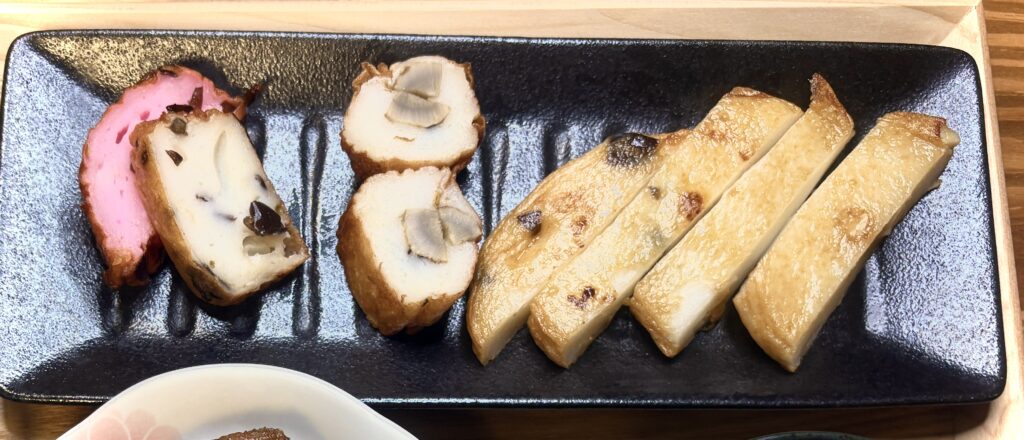 黒い長方形の皿に盛られたさつま揚げの盛り合わせ。左からピンク色が鮮やかな紅しょうが入り、黒きくらげが練り込まれたもの、きのこを包んだ丸いさつま揚げ、香ばしく焼き色がついた長方形のさつま揚げが並んでいる。魚の旨みと具材の食感が楽しめる有名店の一品。