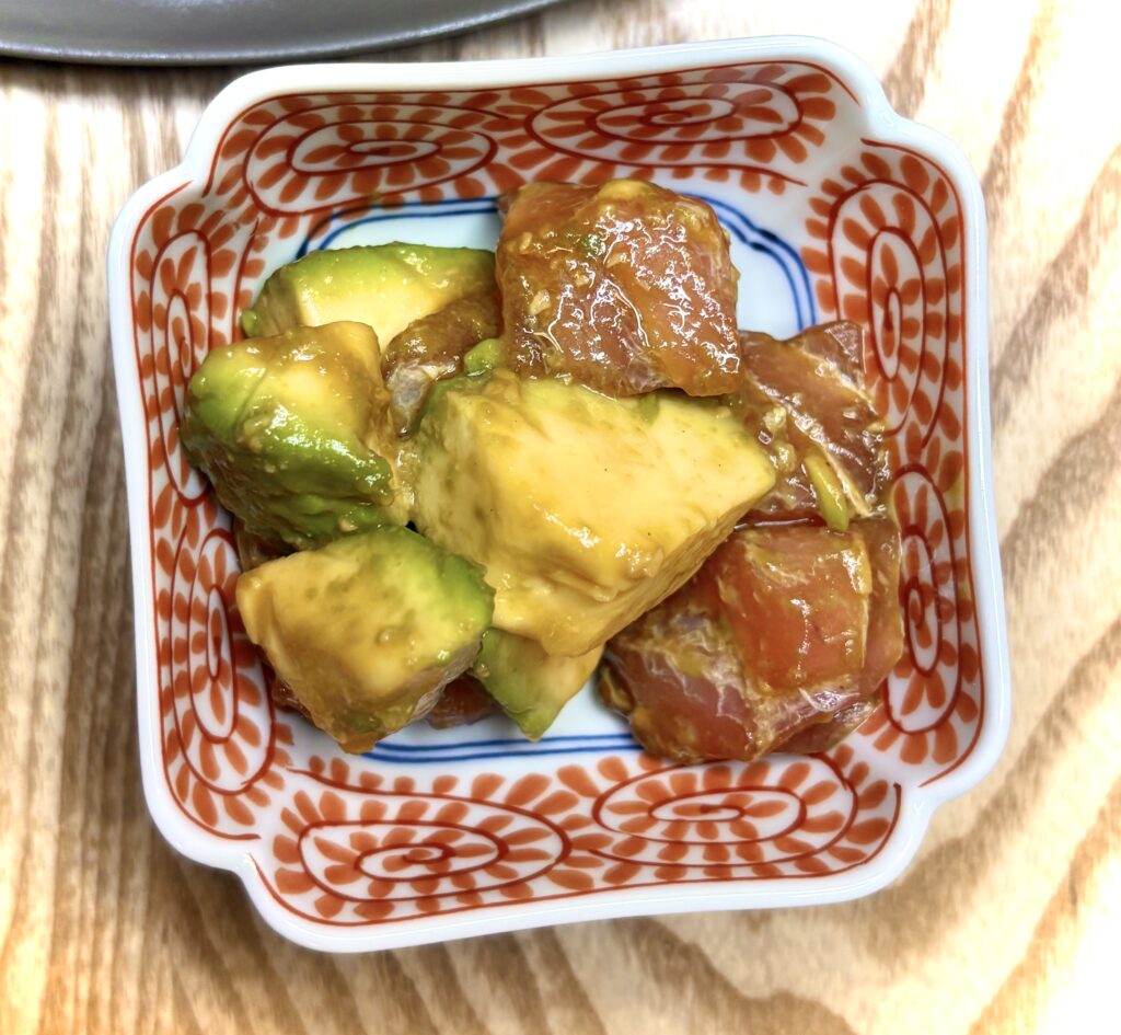 マグロとアボカドの山葵醤油和え。角切りにしたマグロとアボカドをわさび醤油で和えた副菜が、赤い模様の小鉢に盛り付けられている。