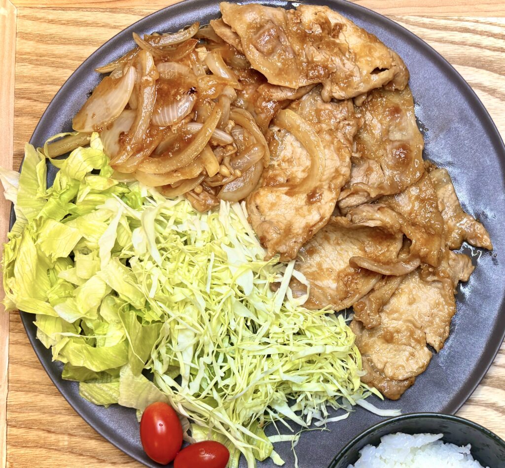 「豚の生姜焼き。玉ねぎと甘辛ダレで炒めた豚肉をレタス、千切りキャベツ、プチトマトと一緒に盛り付けたプレート。」