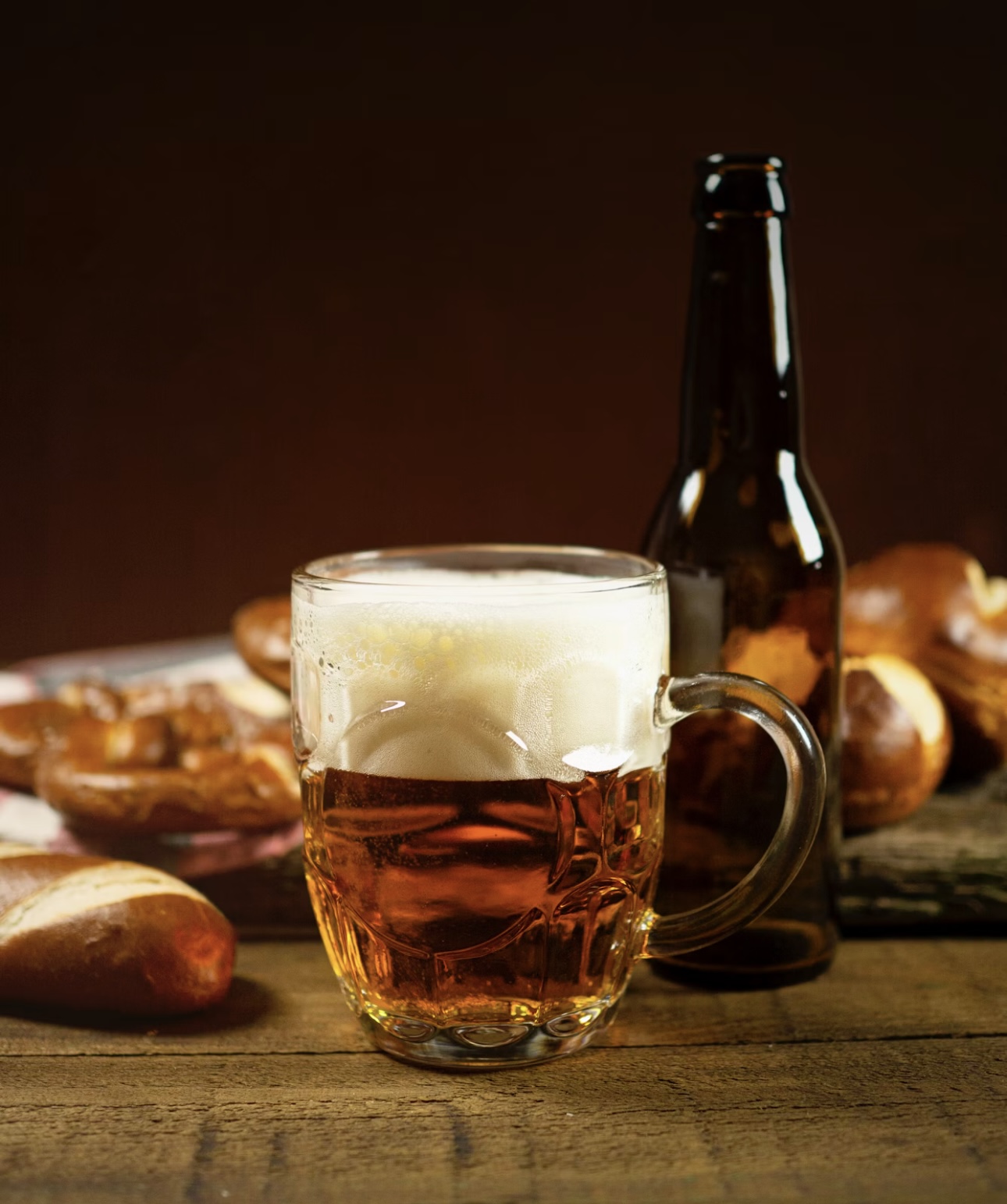 泡がこんもりと立ったジョッキビールと茶色のビール瓶。木のテーブルにプレッツェルが添えられ、温かみのある雰囲気で撮影された一枚。