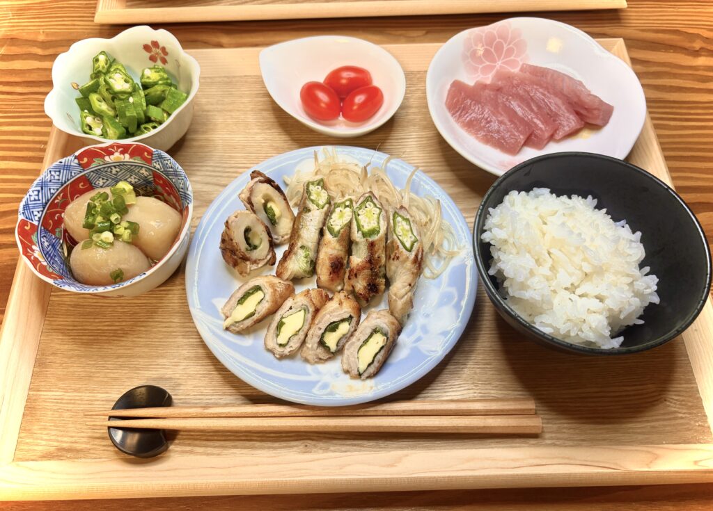 「木のトレーに並んだ和食の定食。メインはオクラやチーズを巻いた豚肉の肉巻き、付け合わせに玉こんにゃくの煮物、刻んだオクラ、ミニトマト、マグロの刺身、そして白ごはんが添えられている。」