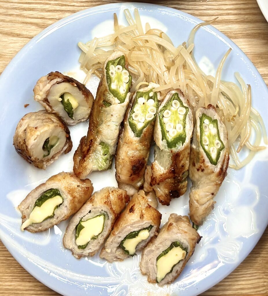 水色の皿に盛られた豚肉の肉巻き。オクラを巻いたものと、チーズと大葉を巻いたものが並び、付け合わせに炒めたもやしが添えられている