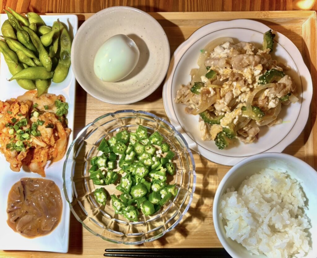 木製トレイに並んだ和風定食。ゴーヤチャンプルー、白ご飯、ゆで卵、枝豆、豚キムチ、イカの塩辛、刻んだオクラの小鉢が彩りよく盛り付けられている。栄養バランスの取れた家庭的な晩ごはん。