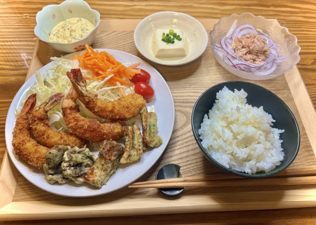 「木のトレーに並んだ和定食。メインの皿にはエビフライや磯辺揚げ、キャベツ・人参・プチトマトのサラダが盛られている。小鉢には豆腐、ツナと紫玉ねぎのサラダ、タルタルソース。右下には白ご飯の茶碗。」