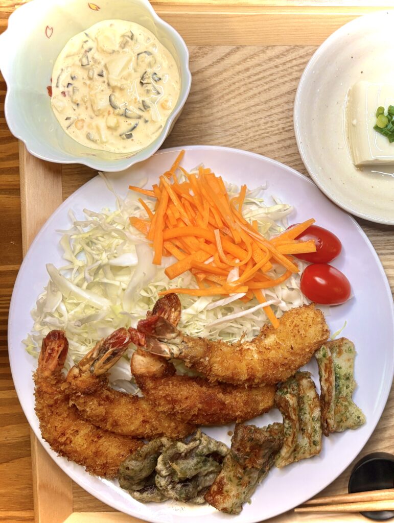 「木製トレーに並んだ和定食。大皿にはエビフライ、磯辺揚げ、千切りキャベツ、千切り人参、プチトマト。小鉢にはタルタルソース、冷奴、ツナと紫玉ねぎのサラダ。右下に白ご飯の茶碗が添えられている。」