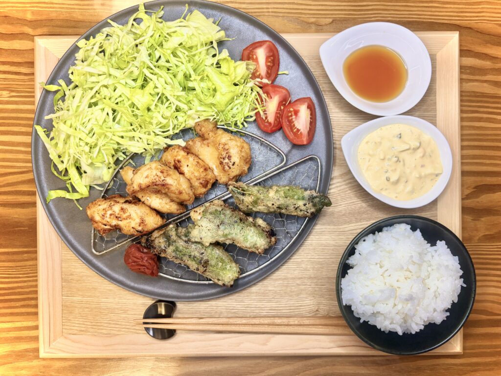 和食の定食セット。メインの皿には揚げたての一口チキン南蛮4個と紫蘇巻き3個が網の上に盛り付けられ、たっぷりの千切りキャベツとカットトマト、梅干しが添えられている。小鉢にはタルタルソースとタレ、手前には白米と箸が並んでいる。
