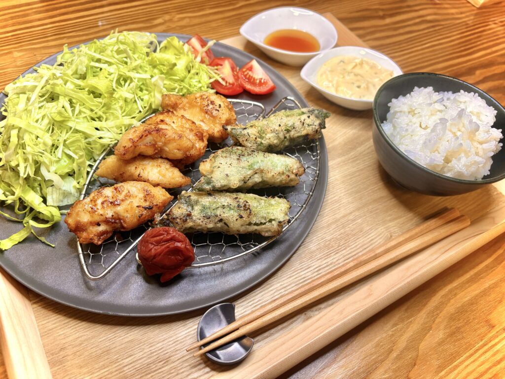 木製トレーに並んだ夕食セット。黒い丸皿に一口サイズのチキン南蛮と紫蘇巻きチキン、キャベツの千切り、トマト、梅干しが盛られている。横には白い器に甘酢とタルタルソース、ご飯茶碗、箸が添えられている。