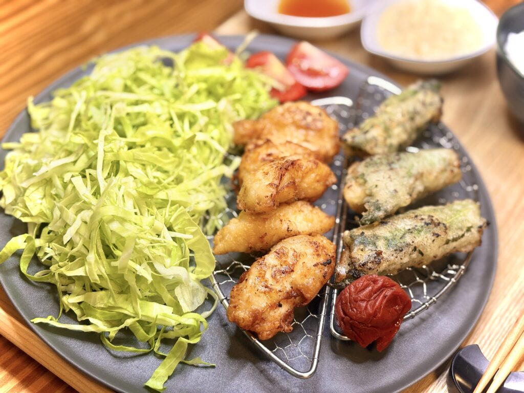 黒い丸皿に盛られた夕食。手前には一口サイズのチキン南蛮、奥には紫蘇で巻いたチキンの揚げ物が並び、横に赤い梅干しが添えられている。左側にはたっぷりのキャベツの千切りとトマトが彩りを添えている。背景には甘酢とタルタルソースの小鉢がぼんやりと見える。