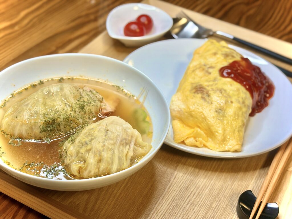 スープでじっくり煮込まれた大きなロールキャベツが二つ並んだ器。隣にはケチャップをのせたオムライスとプチトマトが添えられ、木のトレーにバランスよく盛り付けられている。