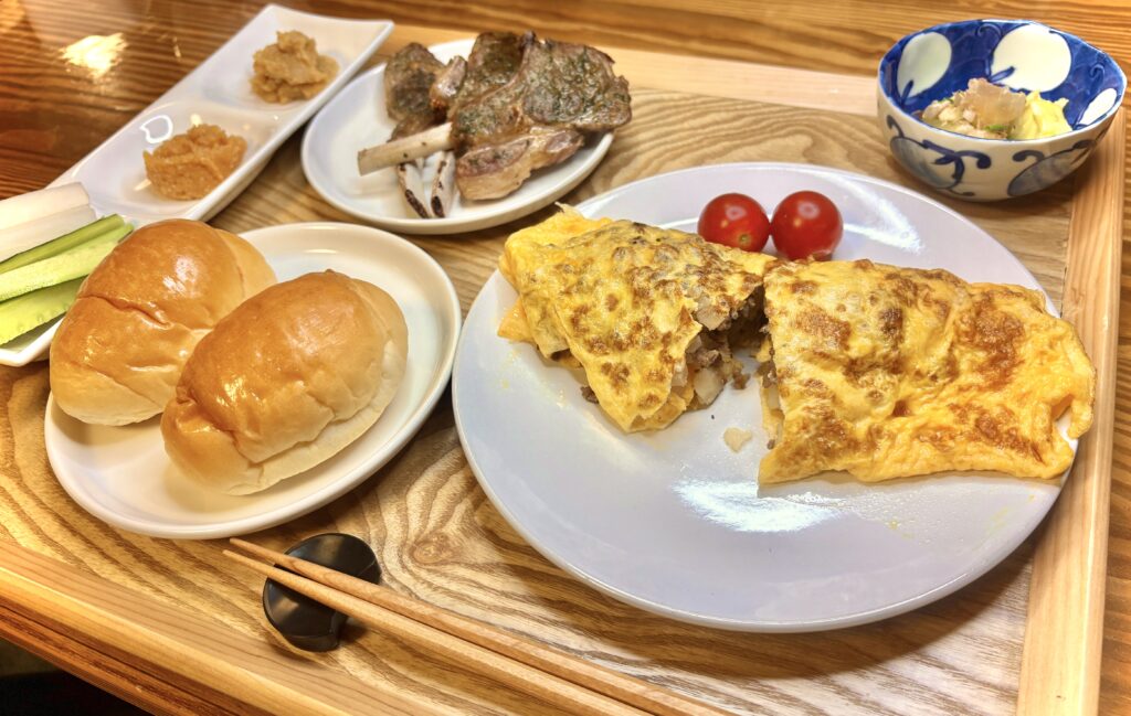 プレートに盛られた夕食セット。具だくさんのオムレツ、ミニトマト、ラムチョップ、焼きなす、パン、野菜スティックと味噌が並び、木のトレイに美しく配置されている。