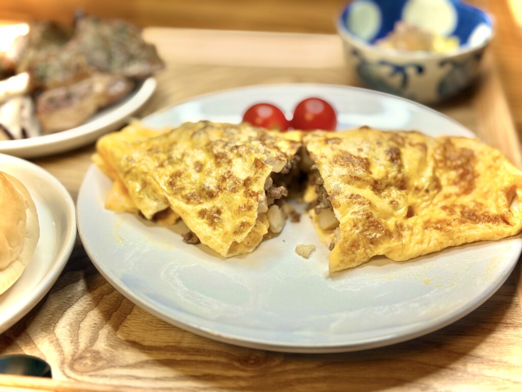 白い皿に盛られた具だくさんのオムレツ。中にはじゃがいもやミンチ肉、玉ねぎが入り、横には赤いミニトマトが添えられている。背景にはラムチョップや焼きなすなどの副菜も写っている。」