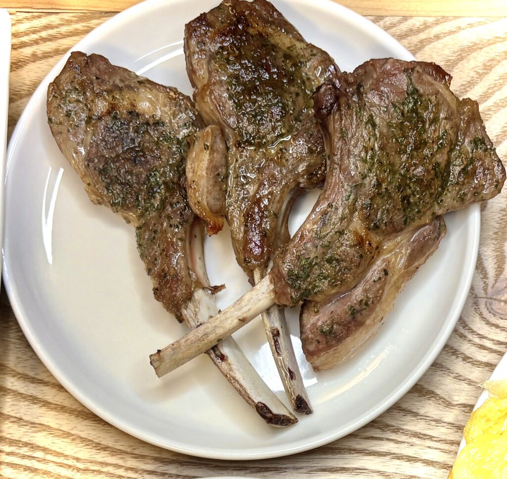 白い皿に盛られた3本のラムチョップ。表面にはこんがりと焼き色がつき、ハーブが散らされて赤ワインとも合う。香ばしく仕上がっている。