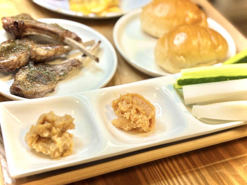 白い仕切り皿に盛られた2種類の味噌と、横に並ぶ大根ときゅうりのスティック野菜。背景にはラムチョップと丸いパンが写っている。