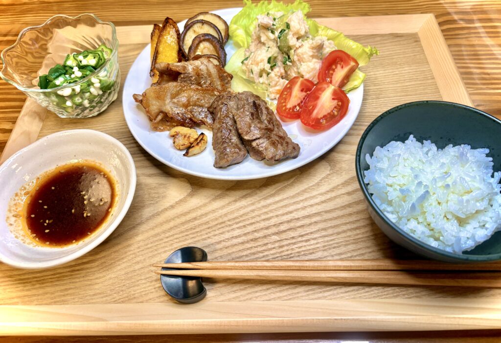木のトレーに盛られた夕食セット。中央の皿には牛肉と豚肉のダブル焼肉、焼きしいたけ、焼きパプリカ、ポテトサラダ、トマトが盛り付けられている。左上には小鉢に刻んだオクラ、左下にはタレの入った小皿、右には白ごはんの茶碗が添えられている。