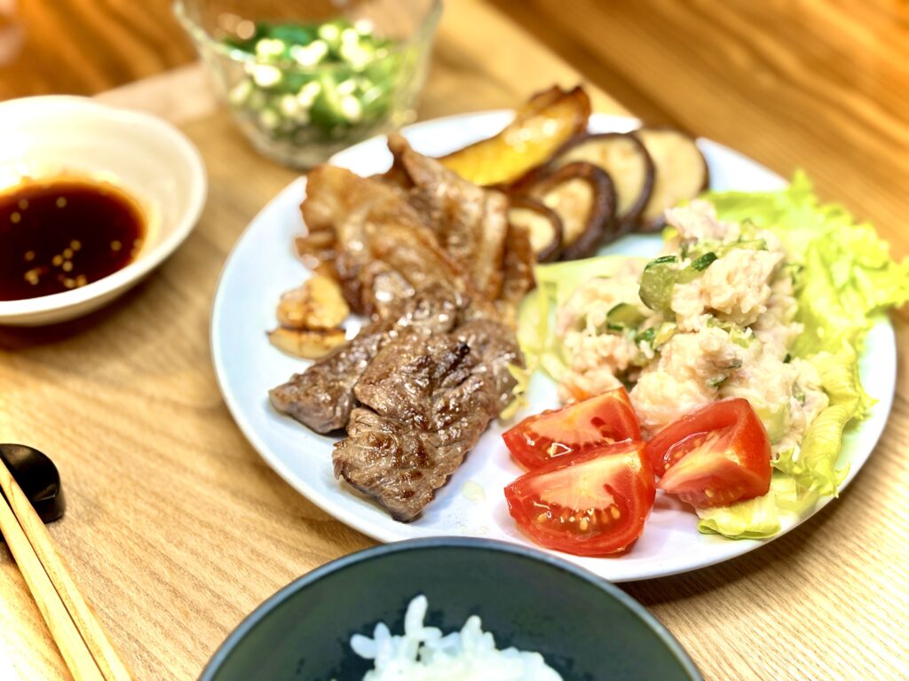 焼肉定食プレート。ジューシーに焼いた牛肉と豚肉、香ばしいパプリカと椎茸のグリル、ポテトサラダとトマト、レタスを盛り付けた彩り豊かなワンプレート。小鉢には刻みオクラ、横には焼肉のタレと白ごはん。