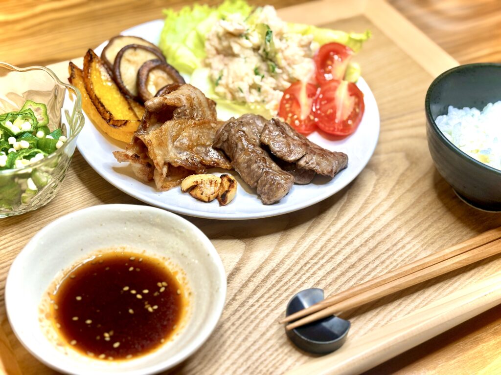 焼肉定食プレート。牛肉と豚肉の焼肉、ガーリックチップ、焼きパプリカと椎茸、ポテトサラダ、トマトとレタスを盛り付けた彩り豊かなワンプレート。小鉢に刻みオクラ、タレの小皿、白ごはんと箸を添えた和食の晩ごはん。
