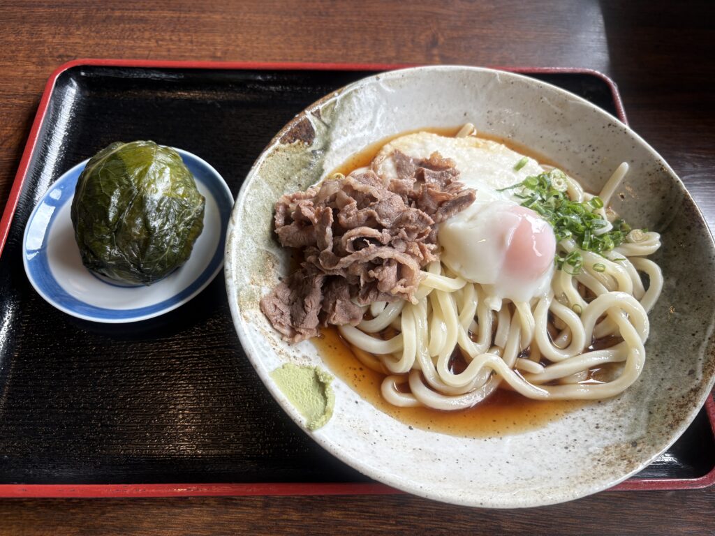 和歌山名物のめはり寿司と、温玉・牛肉・とろろがのったぶっかけうどんのセット。黒いお盆に盛られ、木のテーブルの上に置かれている。