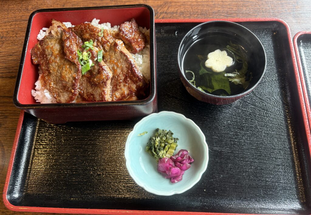 焼肉がのった重箱ご飯と、澄んだお吸い物、漬物のセット。黒いお盆に並べられ、木のテーブルの上に置かれている。