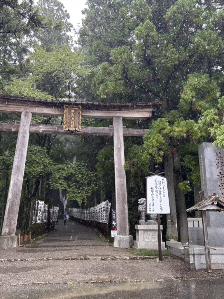 雨の中に立つ熊野本宮大社の大鳥居。両側には石の狛犬と奉納旗が並び、濃い緑の木々に囲まれて神聖な雰囲気が漂っている。