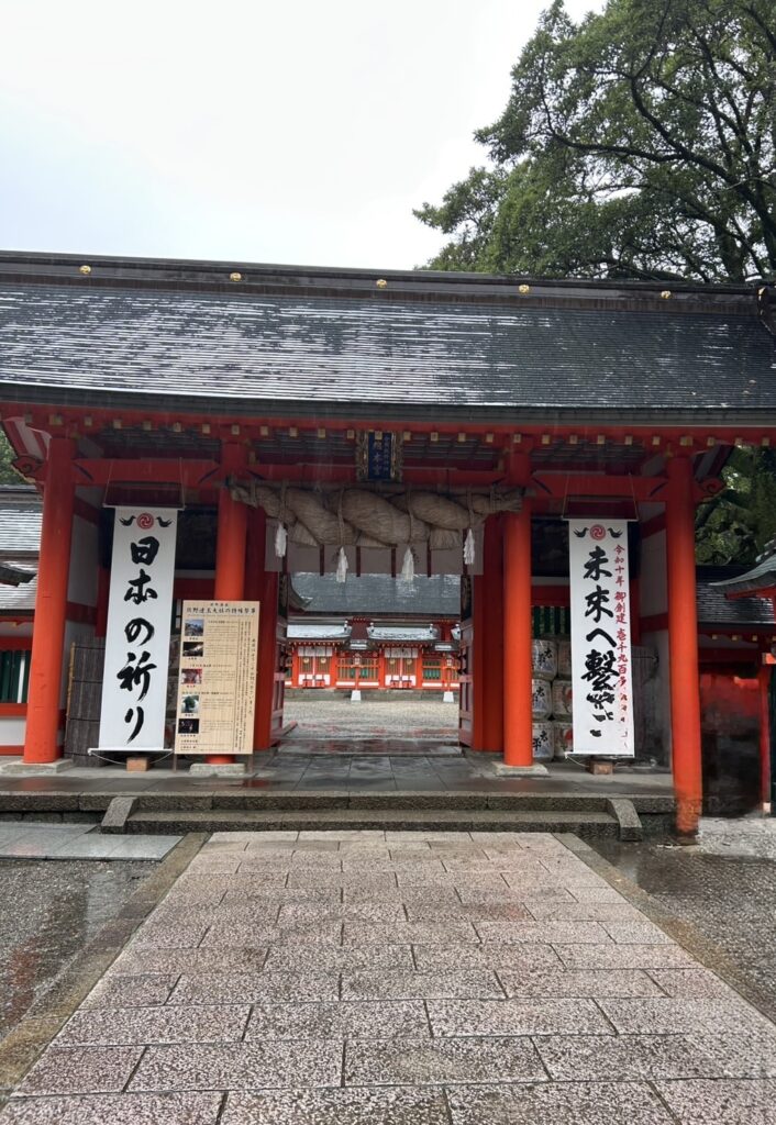 雨に濡れる熊野速玉大社の楼門。赤い柱と大きなしめ縄が特徴で、門の両脇には「日本の祈り」「未来へ繋ぐ」と書かれた大きな看板が掲げられている。