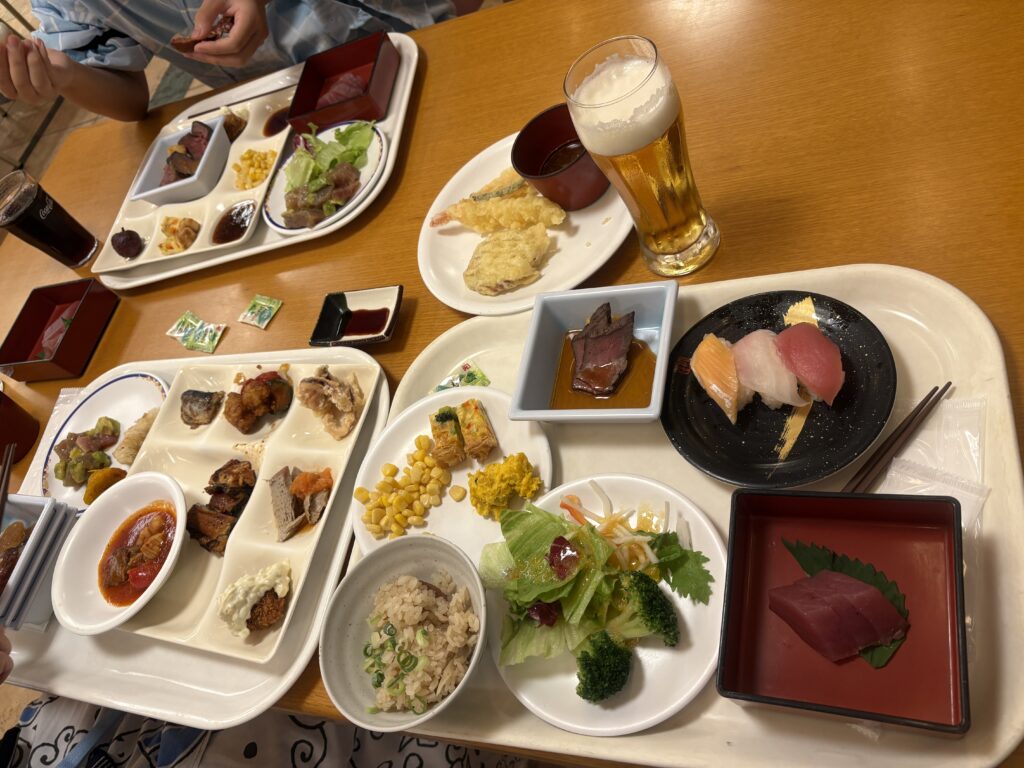 旅館のバイキング料理。寿司、ローストビーフ、天ぷら、煮物、サラダ、炊き込みご飯、デザートなど多彩な料理がトレーに並び、ビールと一緒に楽しめる様子。