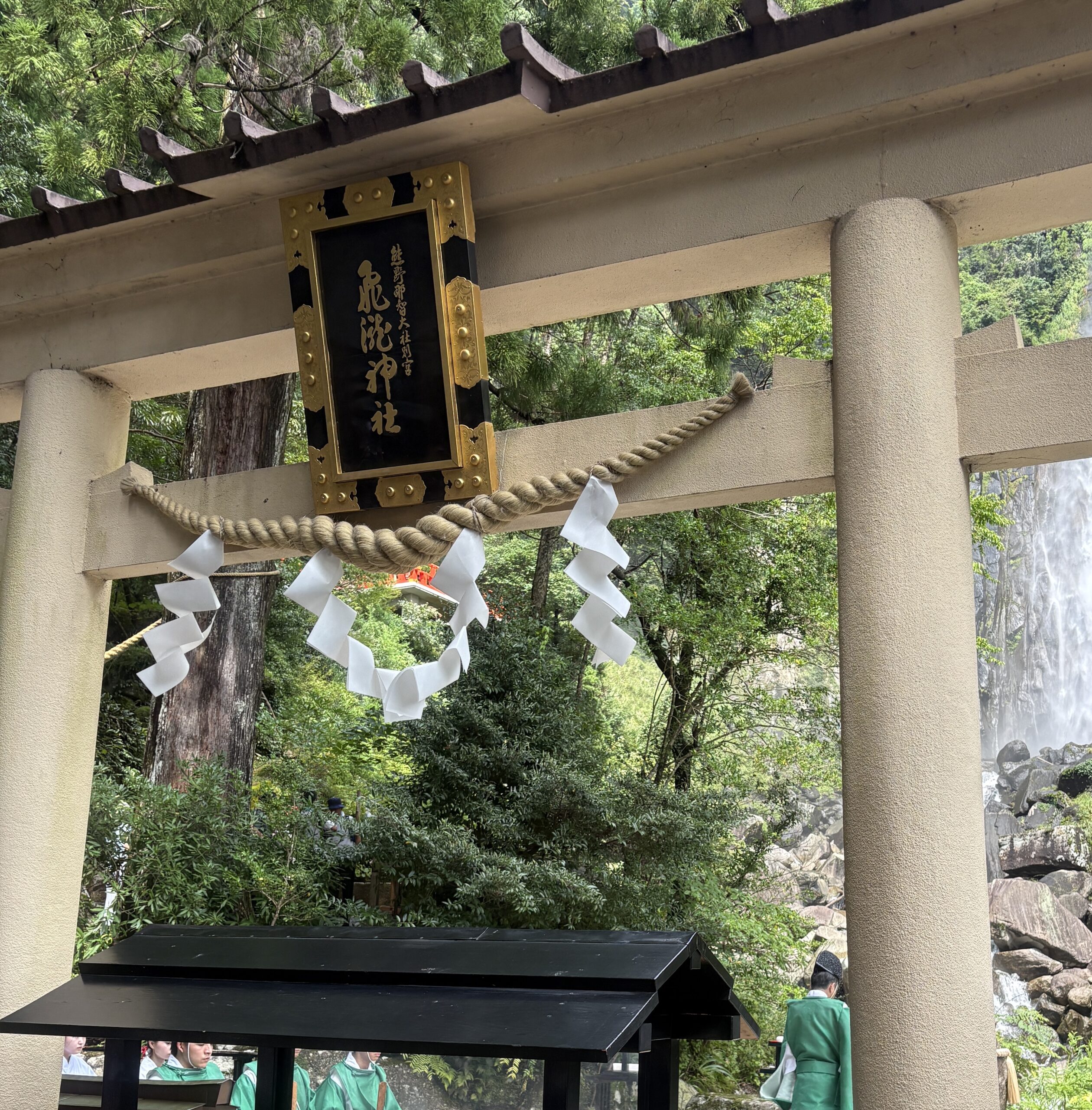 和歌山・那智山にある飛瀧神社の鳥居。しめ縄と紙垂がかかり、奥には緑豊かな森と那智の滝が見える。手前には黒い賽銭箱が置かれ、神職の人々が祭服姿で立っている。