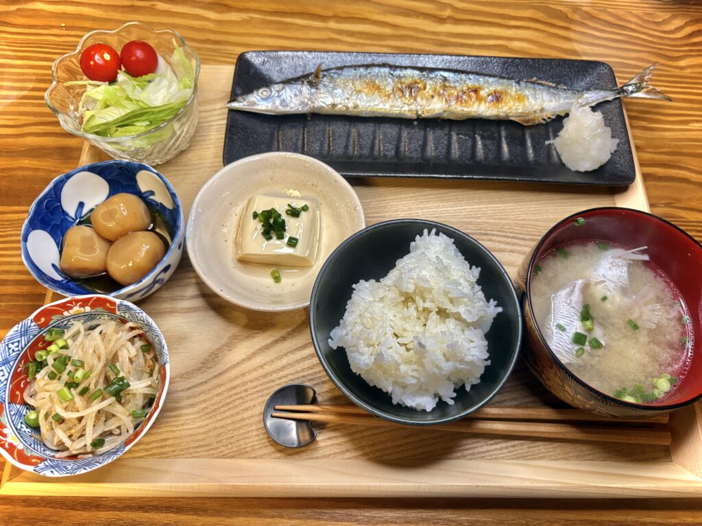 木製のトレイに並んだ和食の献立。焼き魚(さんま)と大根おろし、白ご飯、豆腐の小鉢、里芋の煮物、もやしのナムル、レタスとミニトマトのサラダ、そして具入りの味噌汁。バランスの取れた家庭的な和定食。