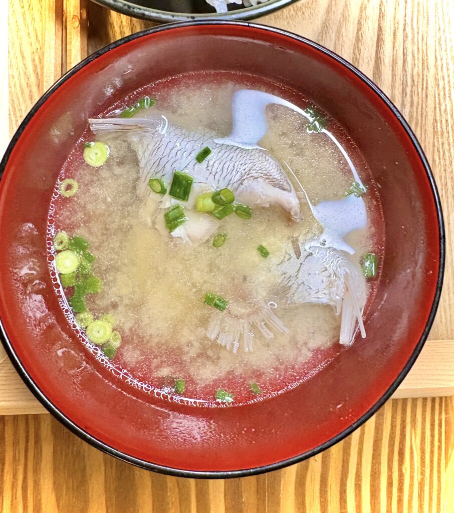 鯛のあらが入った味噌汁。赤いお椀に盛られ、刻みネギが散らされている。鯛の身と骨から出た旨みがたっぷりの和食の一品。