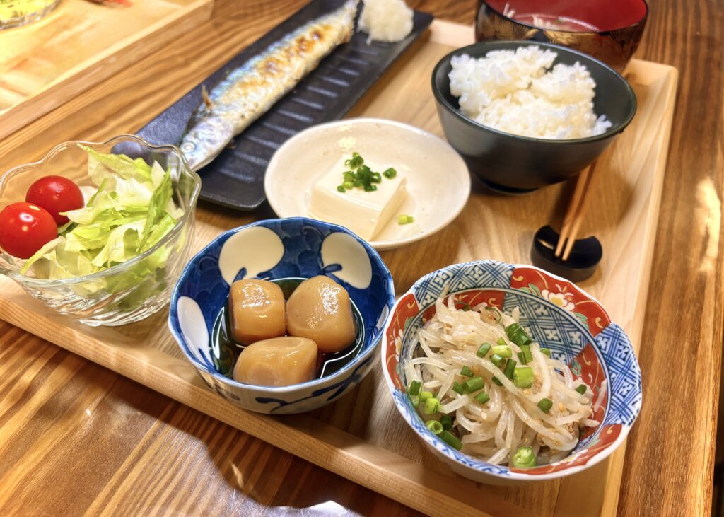 木の盆に並んだ和食の夕食セット。さんまの塩焼き、冷奴、玉こんにゃくの煮物、もやしのナムル、サラダ、ご飯、味噌汁が揃い、彩り豊かで栄養バランスの良い献立。