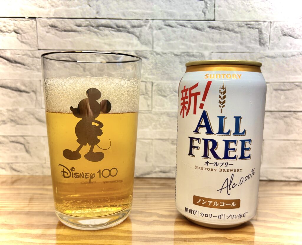 サントリーのノンアルコールビール『オールフリー』の缶と、ミッキーのデザインが入ったグラスに注がれた黄金色の泡立つドリンク。木目調のテーブルと白いレンガ調の壁を背景に撮影
