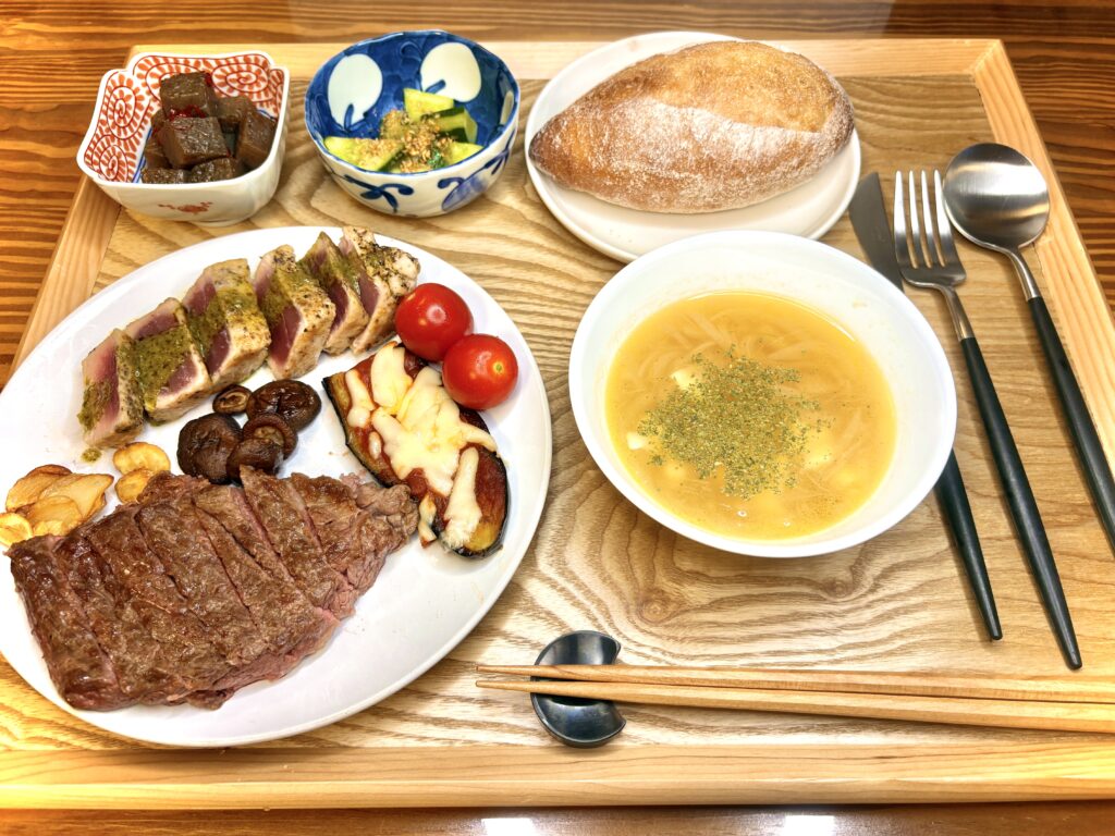 休日の気合いごはん。ステーキ、ビンチョウマグロのソテー(バジルソース)、ナスのピザ風、しいたけとトマト、ピリ辛こんにゃく、きゅうりのナムル、チーズ入りオニオンスープ、人気店のハードパンをワンプレートで並べた豪華な食卓