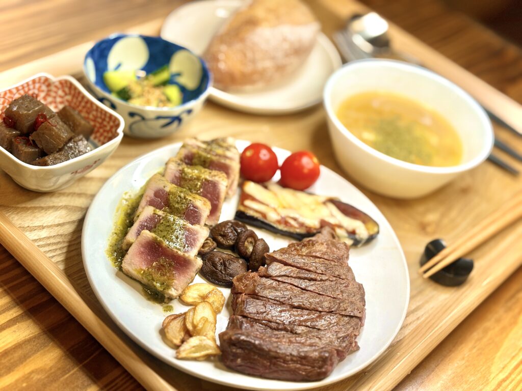 ステーキとビンチョウマグロのソテー(バジルソース)、ナスのピザ風、ガーリックチップやしいたけ、トマトを盛り合わせたプレート。横にはピリ辛こんにゃく、きゅうりのナムル、チーズ入りオニオンスープ、人気店のハードパンを添えた豪華な夕食