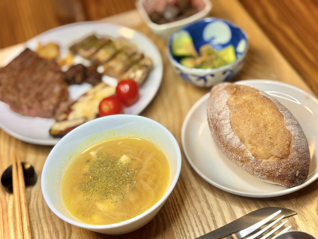 休日の食卓。ステーキとビンチョウマグロのソテー、ナスのピザ風、きゅうりのナムル、ピリ辛こんにゃくに、人気店のハードパンとチーズ入りオニオンスープを添えた豪華なディナー