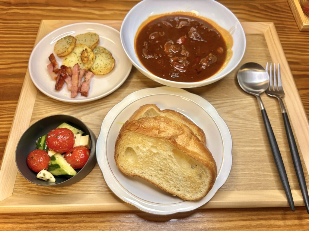木製トレーに並べられた洋風ディナーセット。ゴロゴロ牛肉入りのビーフシチュー、カリッと焼いたジャーマンポテトとベーコン、トマトとチーズときゅうりのカプレーゼ風サラダ、そして厚切りのバゲットが添えられている。スプーンとフォークもセットされ、カフェ風のおしゃれな雰囲気。