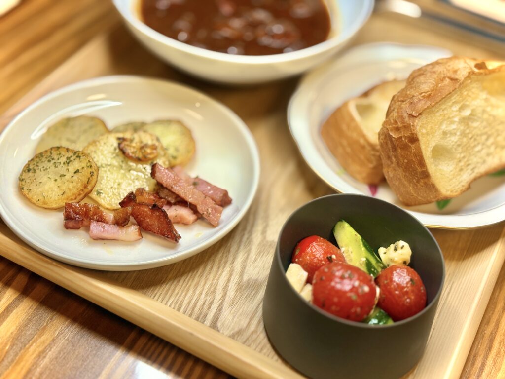 木製トレーに並べられた洋食プレート。手前にはミニトマトときゅうり、チーズを合わせたカプレーゼ風サラダ。奥にはこんがり焼いたジャーマンポテトとベーコン、さらに厚切りのバゲットと濃厚ビーフシチューが添えられている。彩りが良く、カフェ風のおしゃれな雰囲気。