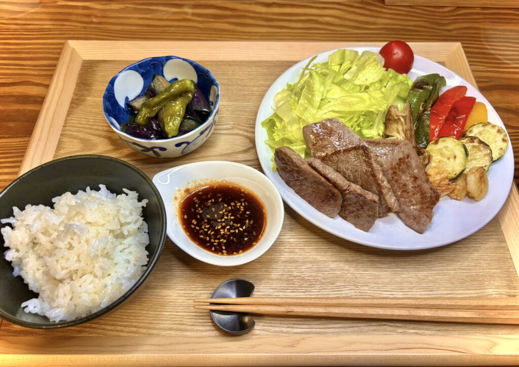 木のトレーに並んだ焼肉定食。厚切りの焼肉と彩り野菜、レタスやミニトマトが盛られた皿に、ご飯、タレ、ししとうとナスの煮浸しの小鉢が添えられている