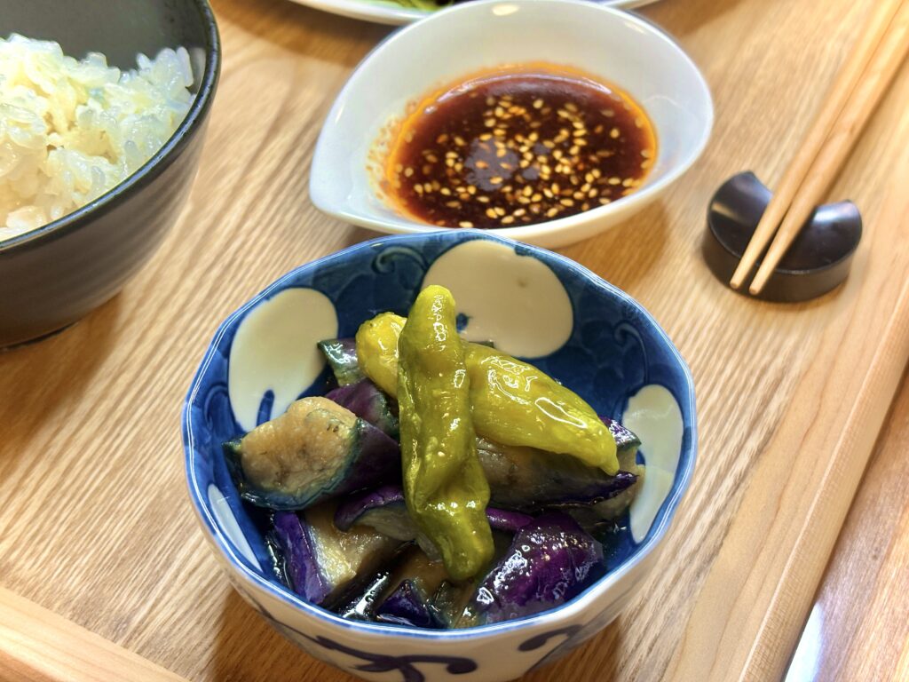 青い模様の小鉢に盛られたししとうとナスの煮浸し。隣には白い器に入ったごま入り焼肉のタレと、ご飯の茶碗、割り箸が添えられている