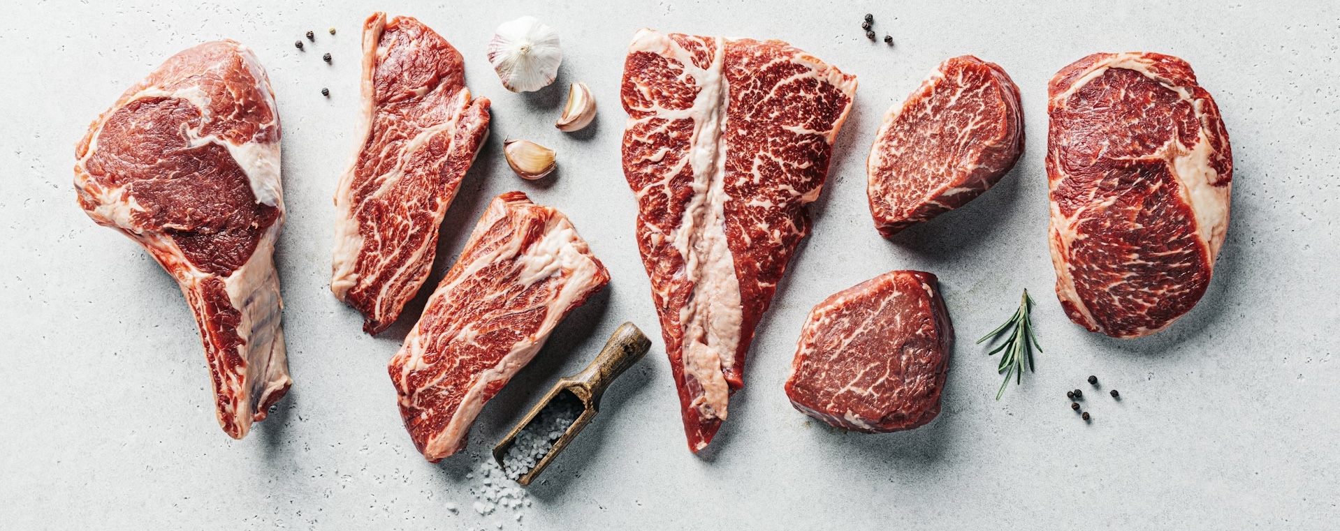 牛肉のさまざまな部位の生肉が並べられた写真。Tボーンステーキやリブ、サーロインなどが置かれ、横にはにんにくやローズマリー、岩塩、黒胡椒が添えられている