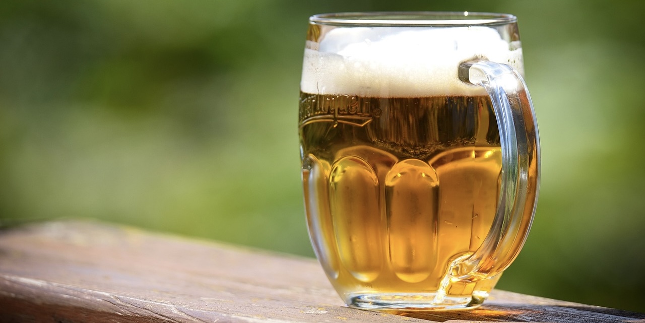 泡が立つ冷えたビールジョッキ。透明なグラスに黄金色のビールが注がれ、屋外の緑を背景に爽やかな雰囲気を演出している