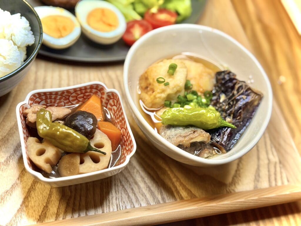 「ナスとししとうが入った揚げ出し豆腐の小鉢と、レンコン・人参・椎茸が入った筑前煮。奥にはご飯と卵、メインプレートが見える」