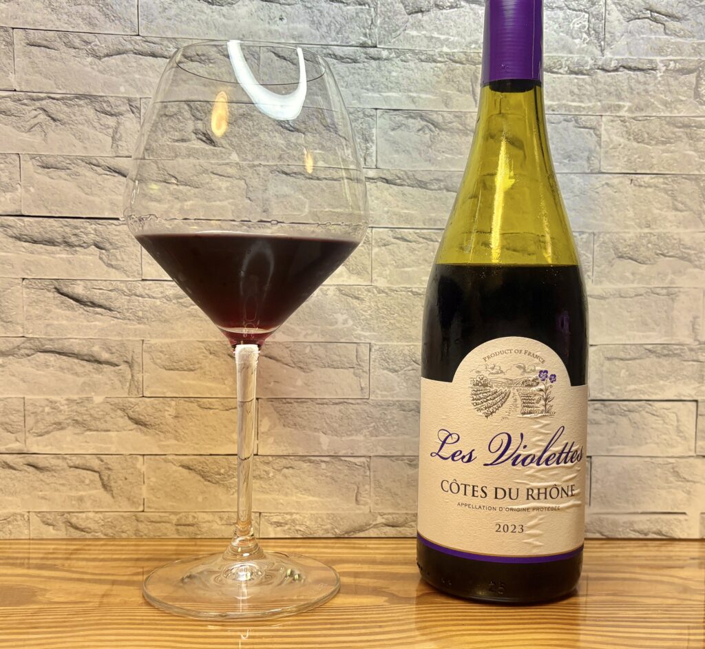 フランス産赤ワイン『Les Violettes Côtes du Rhône 2023』のボトルとワイングラス。グラスには深みのある赤色のワインが注がれており、木目のテーブルに置かれたスタイリッシュな構図