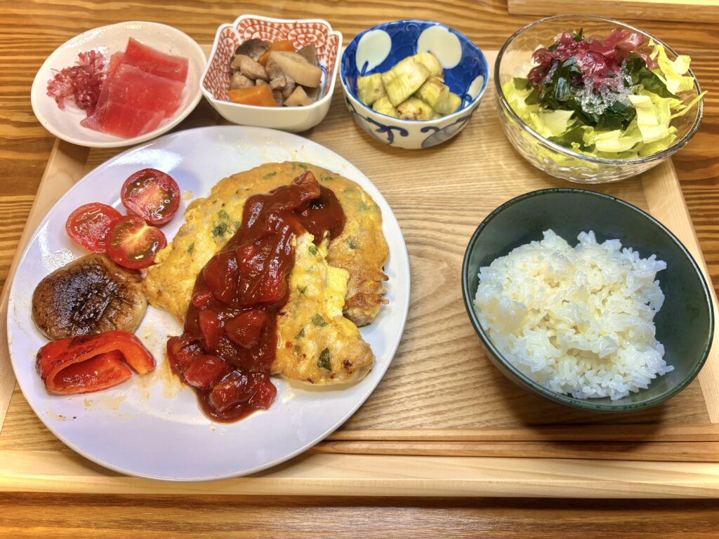 夕食プレート。メインはバジルとチーズのポークピカタにトマトソースをかけた一皿。付け合わせに椎茸と赤パプリカのソテー、プチトマト。小鉢には筑前煮、焼きなす、海藻サラダ、まぐろの切り身。白ごはんを添えた和洋折衷の献立