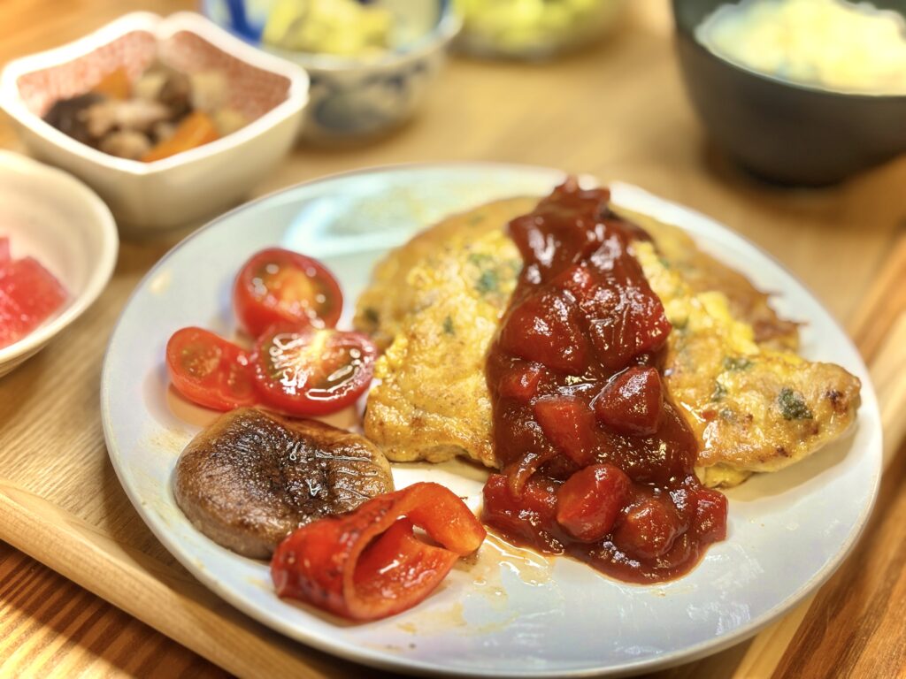 バジルとチーズのポークピカタにトマトソースをかけたメイン料理。付け合わせに焼き椎茸、赤パプリカのソテー、プチトマトを添えた夕食プレート。奥にはまぐろの刺身や筑前煮など小鉢も見える和洋折衷の献立