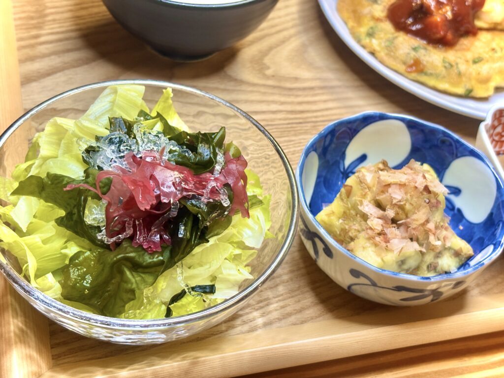 海藻サラダの小鉢。レタスの上にわかめ、赤と透明の海藻が彩りよく盛られている。隣の器には焼きなすを盛り付け、かつお節をトッピングした副菜。和食らしいさっぱりとした小鉢料理」