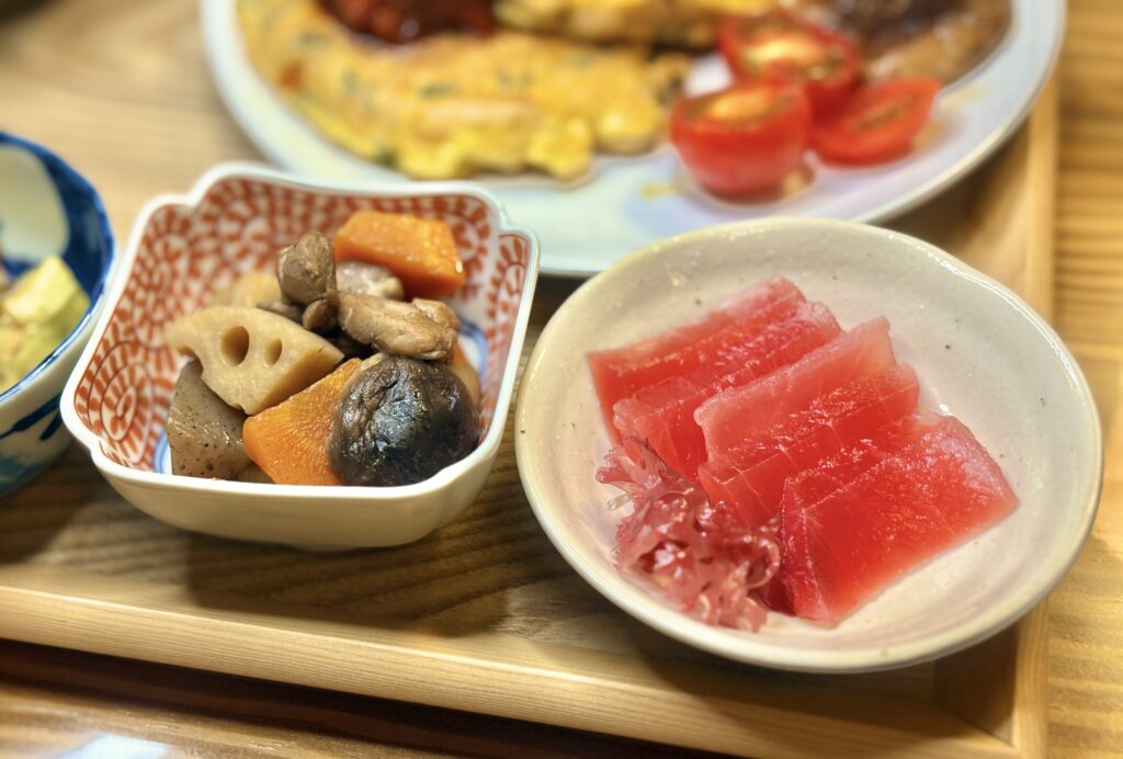 まぐろの刺身を盛り付けた小鉢と、筑前煮の小鉢。筑前煮にはレンコン、にんじん、椎茸、こんにゃく、鶏肉などが彩りよく入っている。奥にはプチトマトを添えた料理も見える夕食の一部