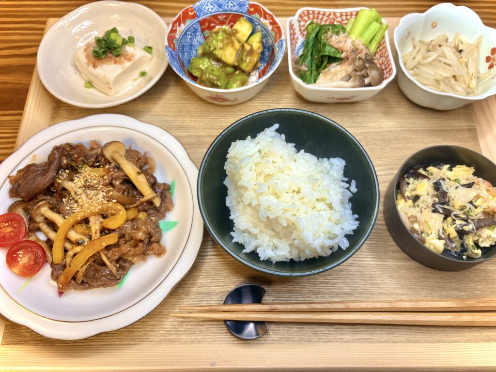 木製トレーに並んだ“おうち居酒屋”風の夕食。メインは牛肉ときのこのプルコギ、彩りのミニトマト添え。中央に白ご飯、右下にはカニカマときくらげの卵とじ。上段には冷奴、アボカドの和え物、小松菜としめじのおひたし、もやしナムルが並び、小鉢が充実した大人の晩酌セットのような食卓。