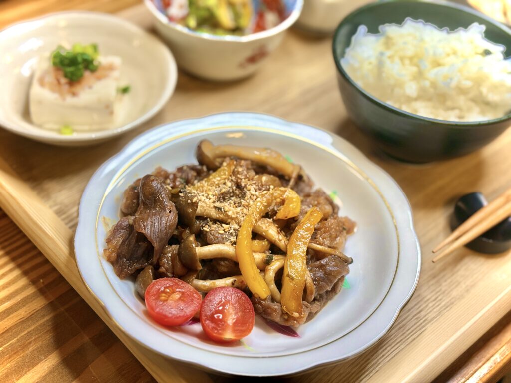 木のトレーに並んだ夕食のアップ。メインの牛肉ときのこのプルコギに黄パプリカとごまを添え、横にプチトマトが彩りを加えている。奥には冷奴や小鉢料理、ご飯が並び、居酒屋風の晩酌セットを思わせる食卓。